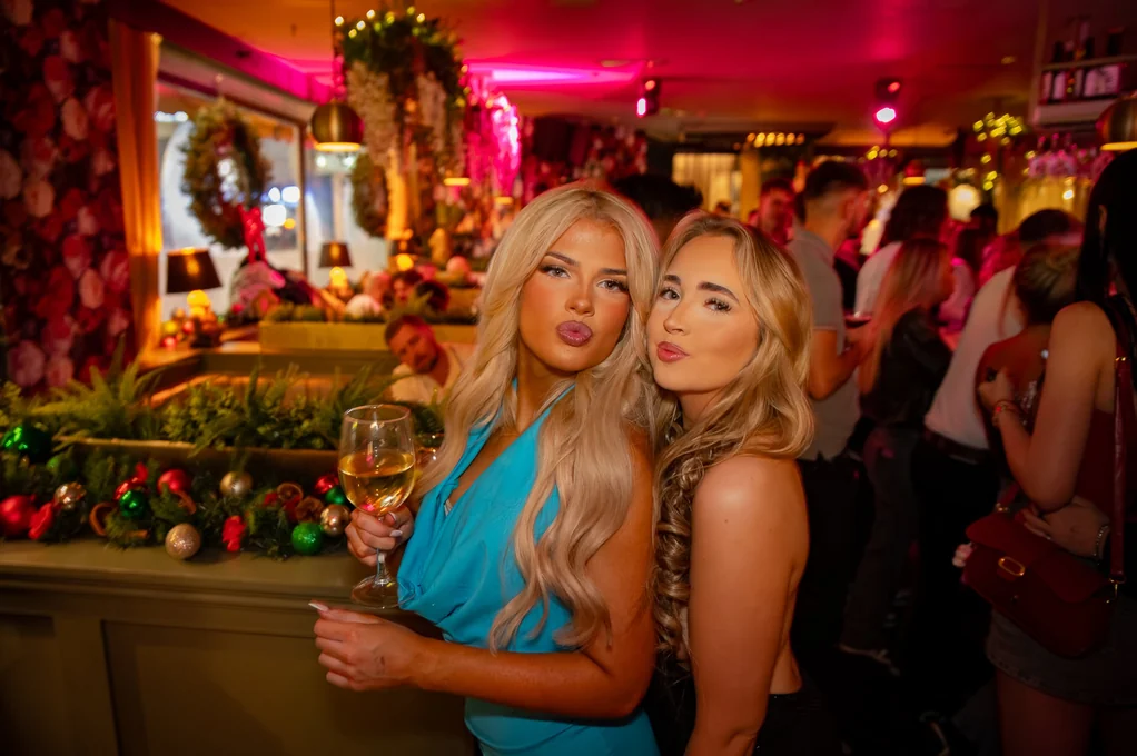 Two friends with drinks at the bar under red and pink Christmas lights