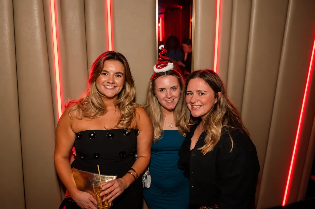 Three friends with drinks in front of the red neon, one in a Santa hat