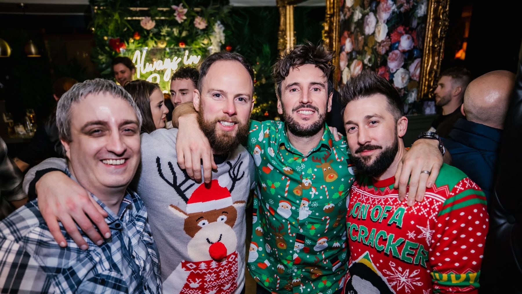 Four friends in Christmas jumpers at a Baluga Christmas party, beneath the tree and the Always in Baluga neon